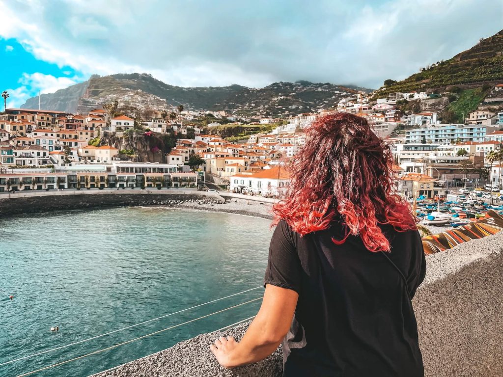Eine Frau mit roten Locken von hinten, an ein Steingeländer gelehnt, mit Blick auf die Bucht von Câmara de Lobos mit bunten Häusern am Hang und türkisfarbenem Wasser