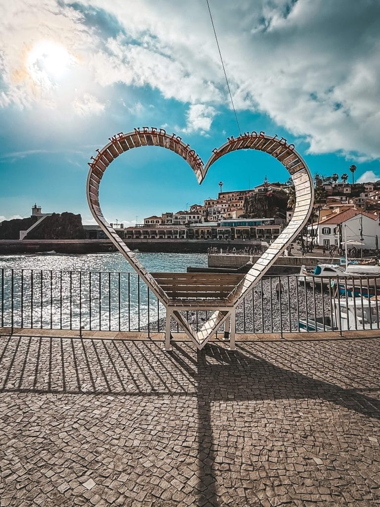 Eine herzförmige Metallskulptur mit dem Schriftzug „All we need is love“ auf Steinpflaster über dem Meer, mit Blick auf die Bucht und die Häuser von Câmara de Lobos, im Gegenlicht.