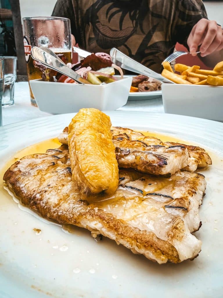 Ein Teller mit gegrilltem Fischfilet, darauf ein Stück gebratene Banane in Buttersoße; im Hintergrund ein zweiter Teller mit Espetada, Pommes und ein Coral-Bier.