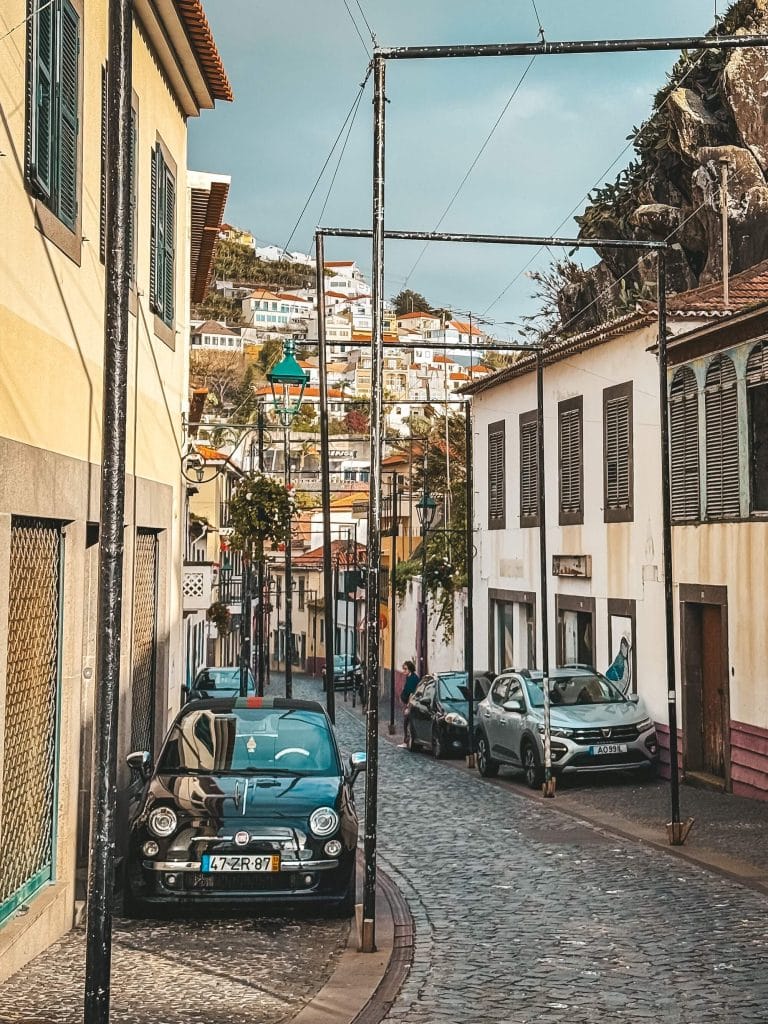 Eine schmale Gasse mit Kopfsteinpflaster und bunten Häusern auf beiden Seiten, ein am Rand geparkter Fiat 500, Kabel zwischen den Häusern, dahinter Gebäude am Hang