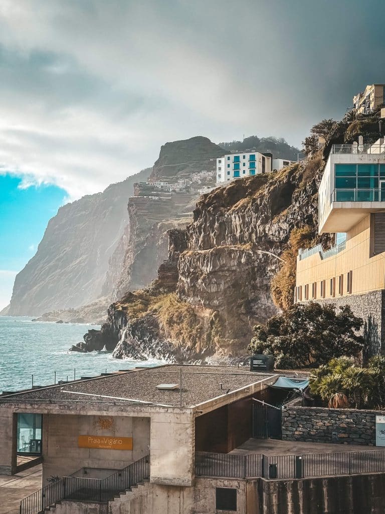 Felsklippen mit Gebäuden direkt an der Kante (darunter Praia do Vigário und ein modernes Hotel), links der Ozean, dahinter verschwindet das mächtige Cabo Girão im Dunst.
