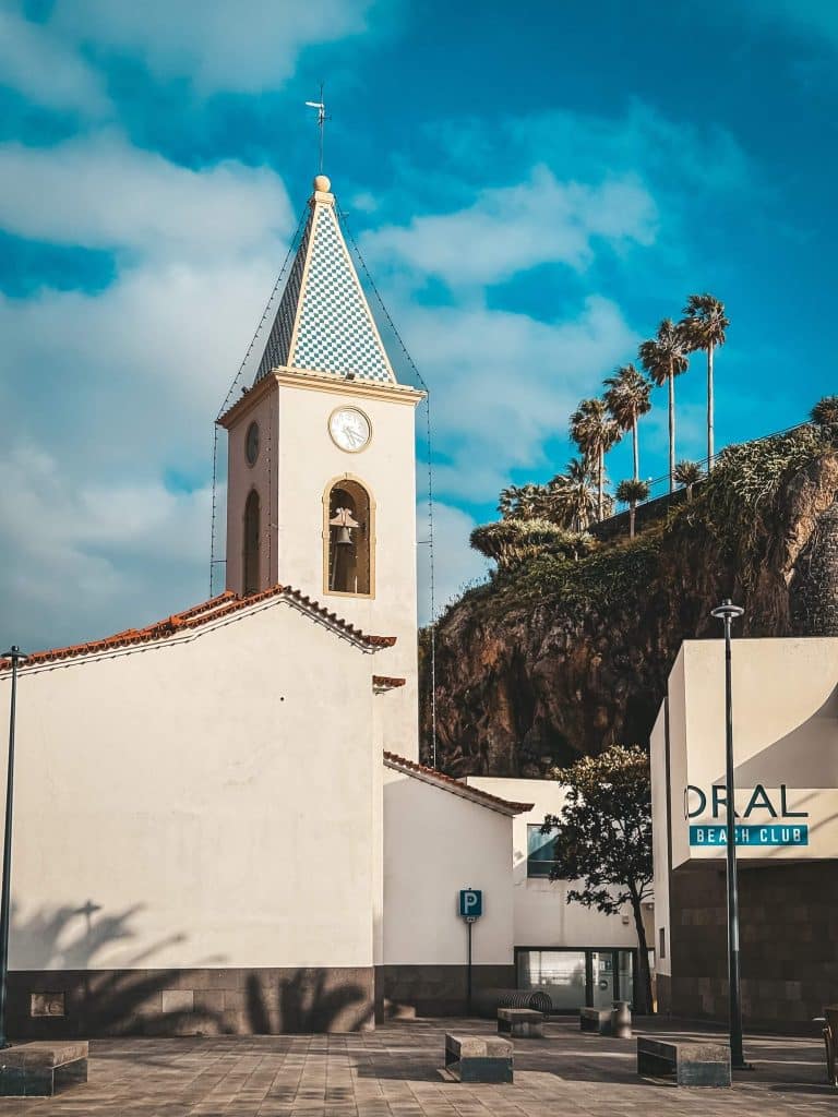 Eine weiße Kirche mit Uhr und einem Turm mit grün-weißem Schachbrettmuster, rechts Palmen und das moderne Gebäude des Coral Beach Club, vor einer Felsklippe.
