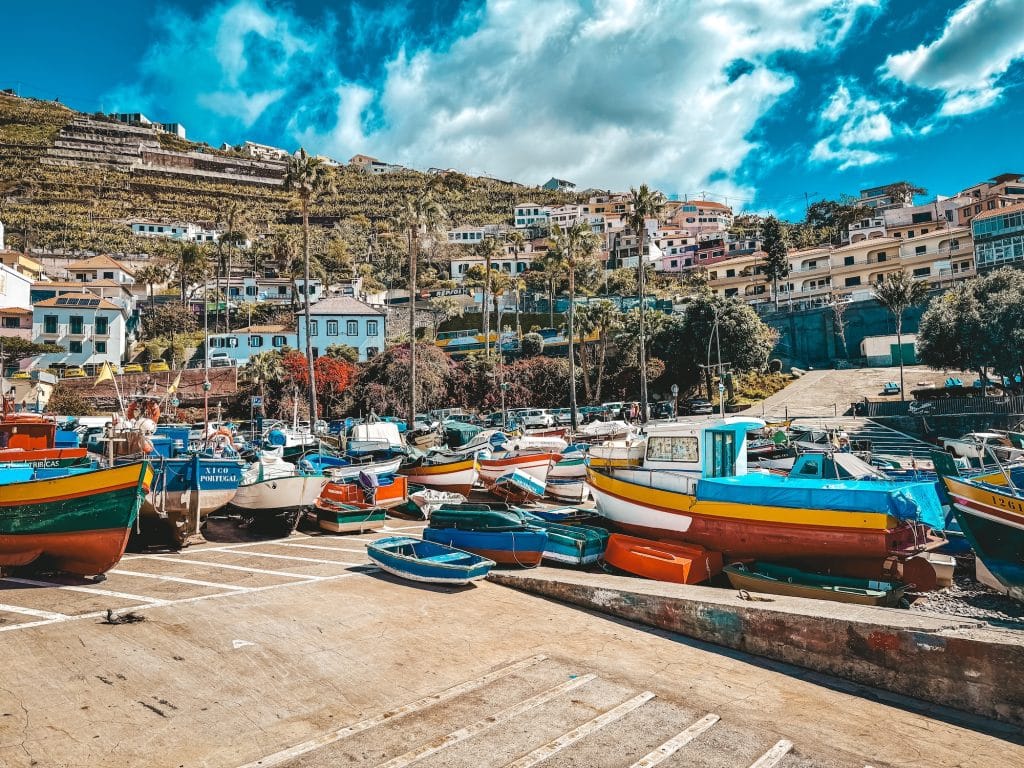 Weitwinkelaufnahme des Hafens von Câmara de Lobos mit Dutzenden bunten Fischerbooten auf einem Betonkai, Weinterrassen an einem steilen Hang und bunten Häusern übereinander am Hügel unter dramatischem Wolkenhimmel.