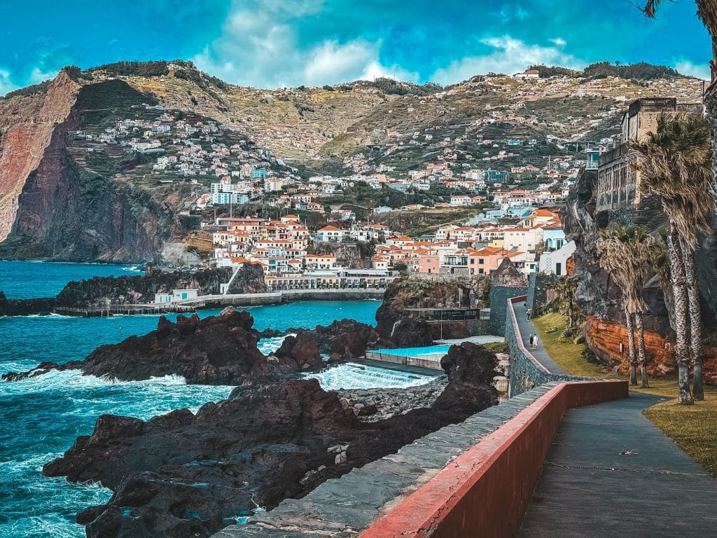 Blick von der Strandpromenade mit rotem Geländer auf dunkle Lavafelsen, die aus dem türkisfarbenen Ozean ragen, natürliche Badebecken und die bunten Häuser von Câmara de Lobos, die sich unter einer mächtigen Klippe den Hang hinaufziehen.