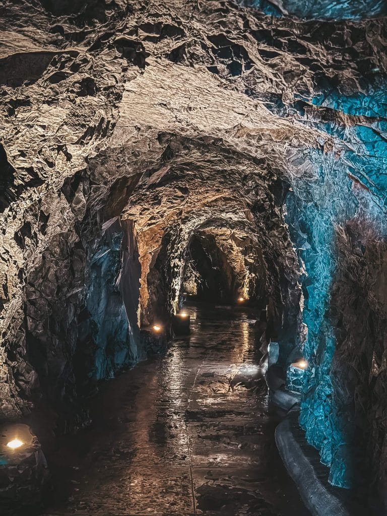 Im Inneren eines Felstunnels mit warmen und blauen Lampen, ein nasser Weg führt hinein, an der Wand entlang verläuft eine Levada.