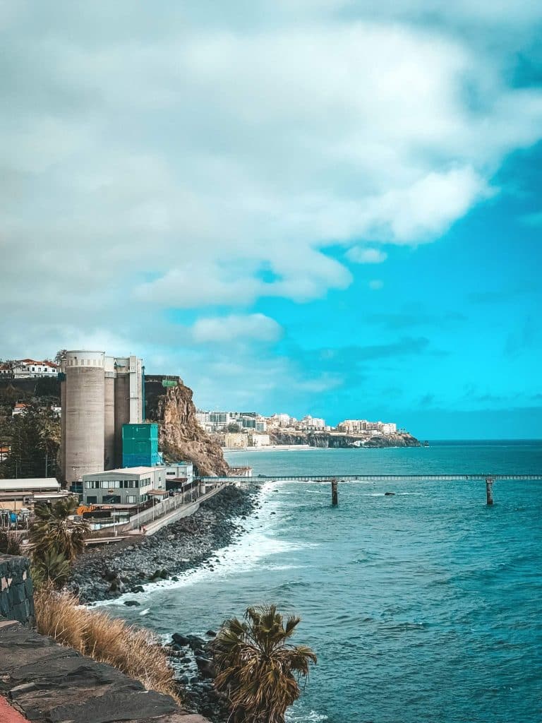 Das Ufer mit Betonsilos und Industriegebäuden links, einem steinigen Strand und einer Brücke auf Pfählen über dem türkisfarbenen Ozean, dahinter die Stadt auf der Klippe.