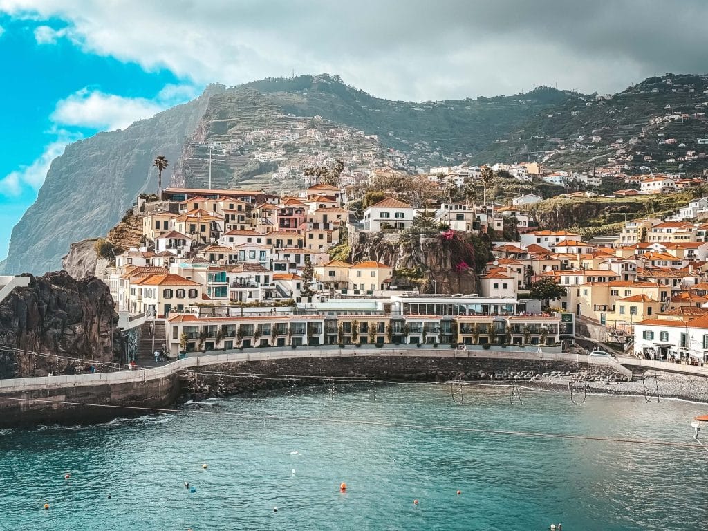 Ein weites Panorama der Bucht von Câmara de Lobos mit türkisfarbenem Wasser, einem Kieselstrand, einer Häuserzeile mit Restaurants am Wasser und bunten Häusern an einem steilen Hang, dahinter eine riesige Klippe.