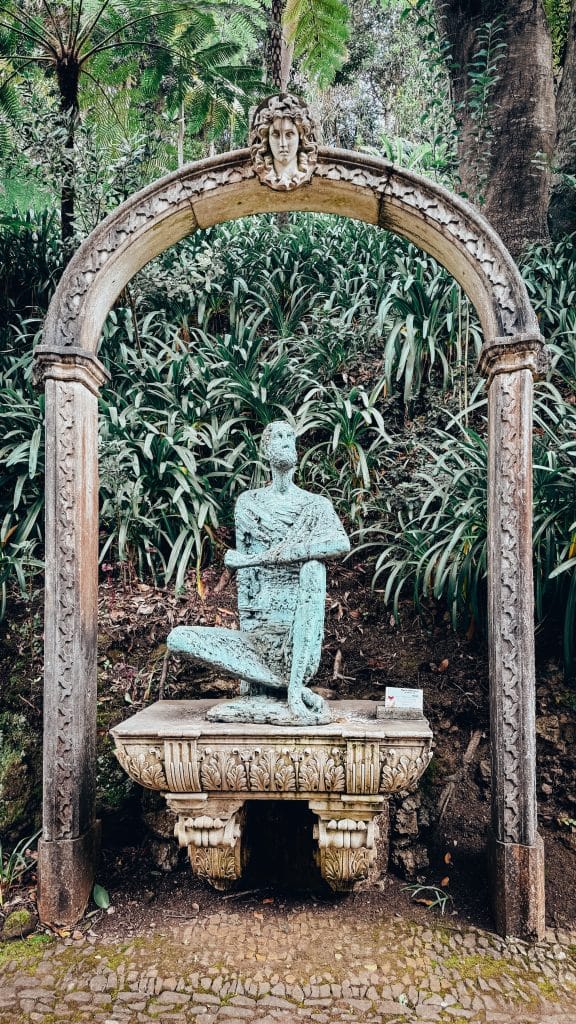 Eine patinierte Bronzeskulptur unter einem Steinbogen zwischen üppigen tropischen Pflanzen im Monte Palace. Foto: Ewelina Podrez-Siama 2025