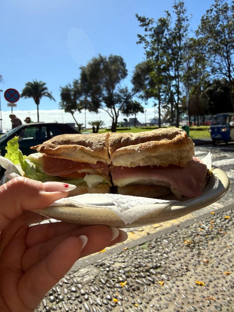 Eine Hand hält einen Teller mit einem Sandwich in einem runden Brötchen mit Fleisch und Salat, dahinter Palmen und Bäume