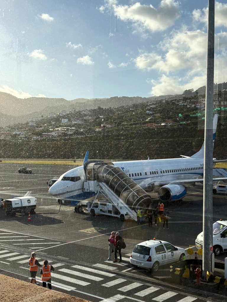Weiß-blaues Flugzeug mit Passagiertreppe auf dem Vorfeld, dahinter ein grüner Hang mit Häusern