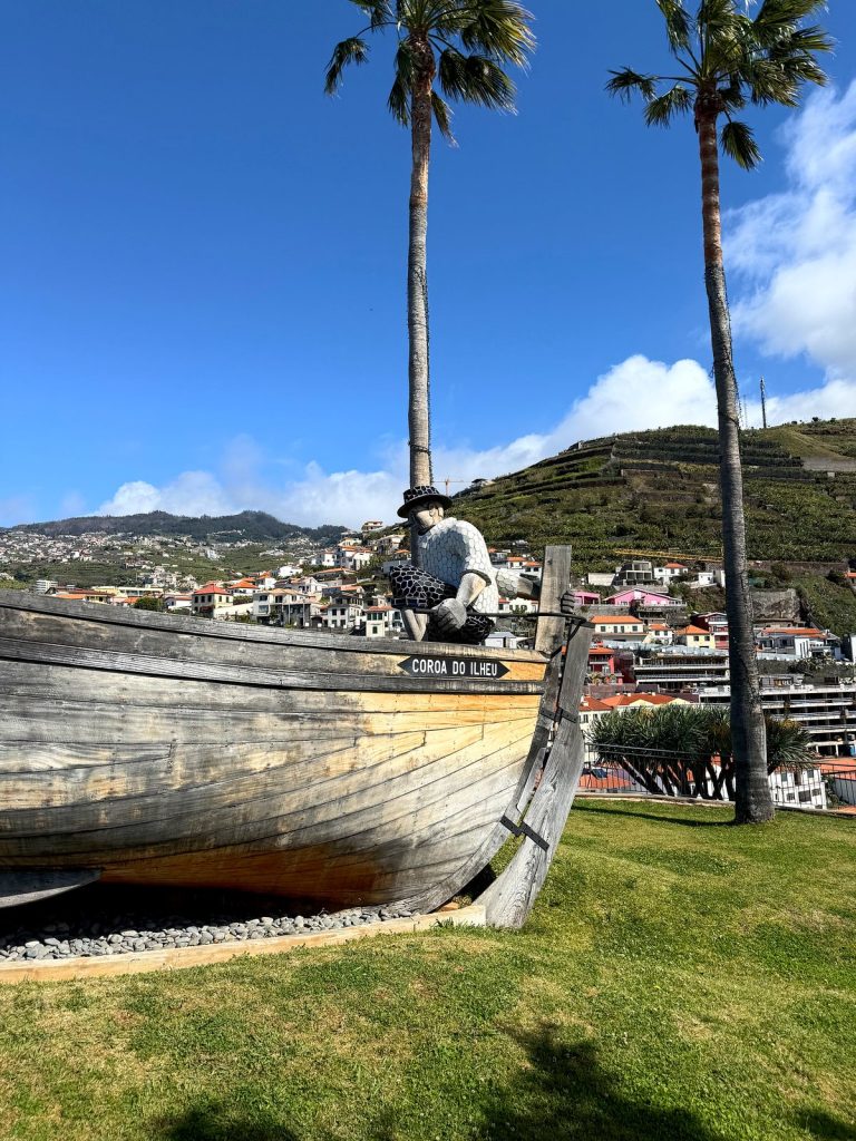 Ein hölzernes Fischerboot mit dem Namen „Coroa do Ilhéu“ als Denkmal auf einer Rasenfläche, am Bug die Figur eines Fischers mit Hut, dahinter Palmen und Häuser von Câmara de Lobos