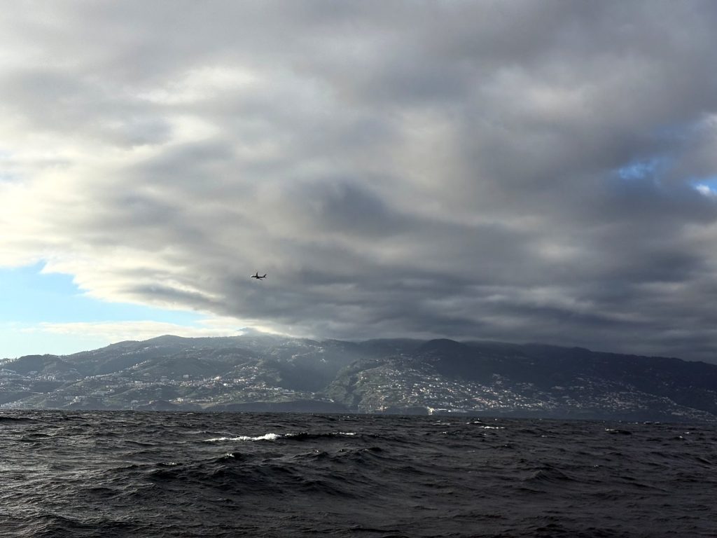 Eine dicke graue Wolkendecke über den Hängen der Küste Madeiras, vom Meer aus gesehen, am Himmel ein Flugzeug