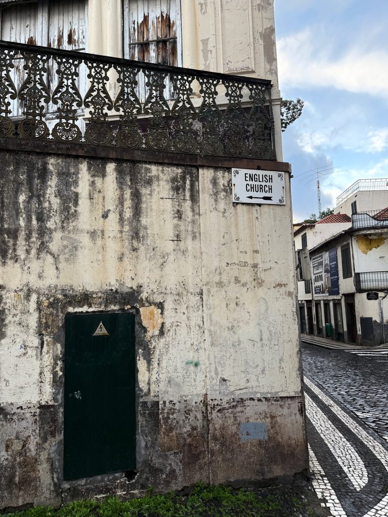 Die abblätternde Wand eines alten Stadthauses mit schmiedeeisernem Balkon und grünen Türen, an der Ecke ein weißes Schild zur English Church, daneben eine gepflasterte Gasse