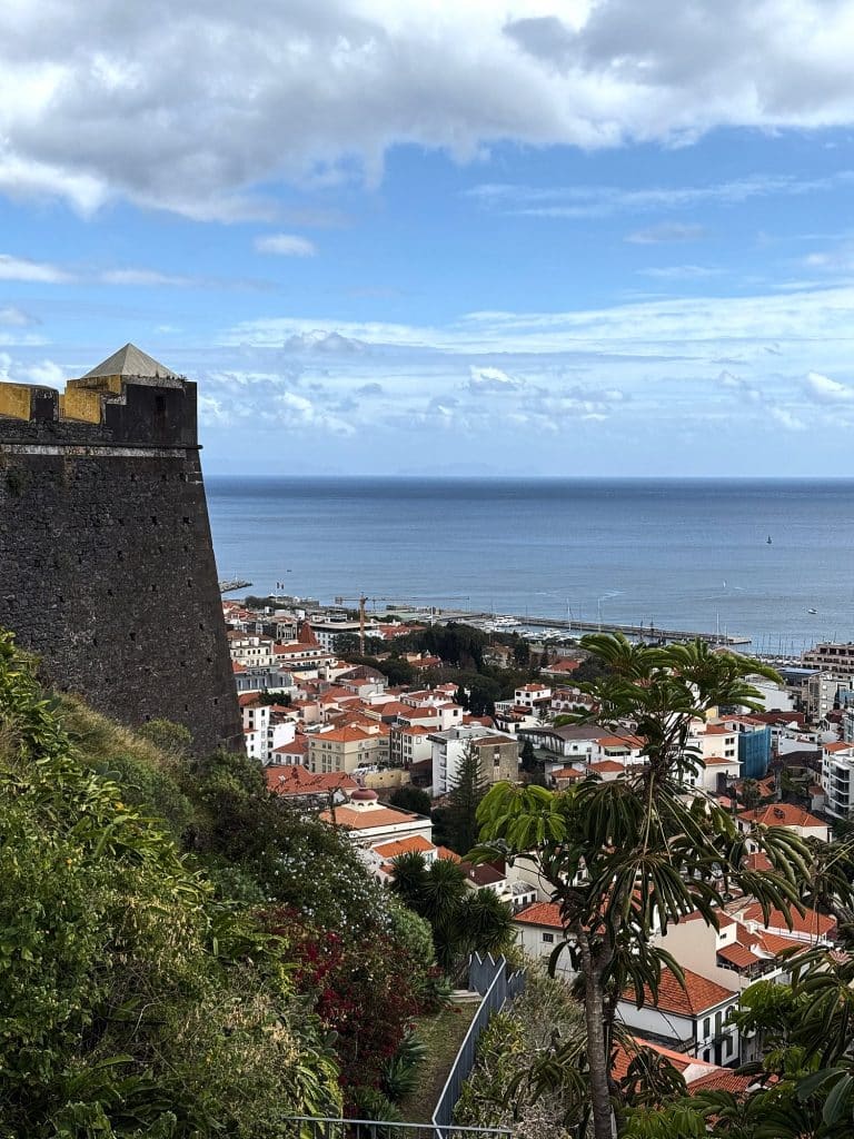 Eckwachturm der Fortaleza do Pico mit Pyramidendach über einem grünen Hang, darunter die Dächer des Zentrums von Funchal und die Hafenmole vor dem Meer