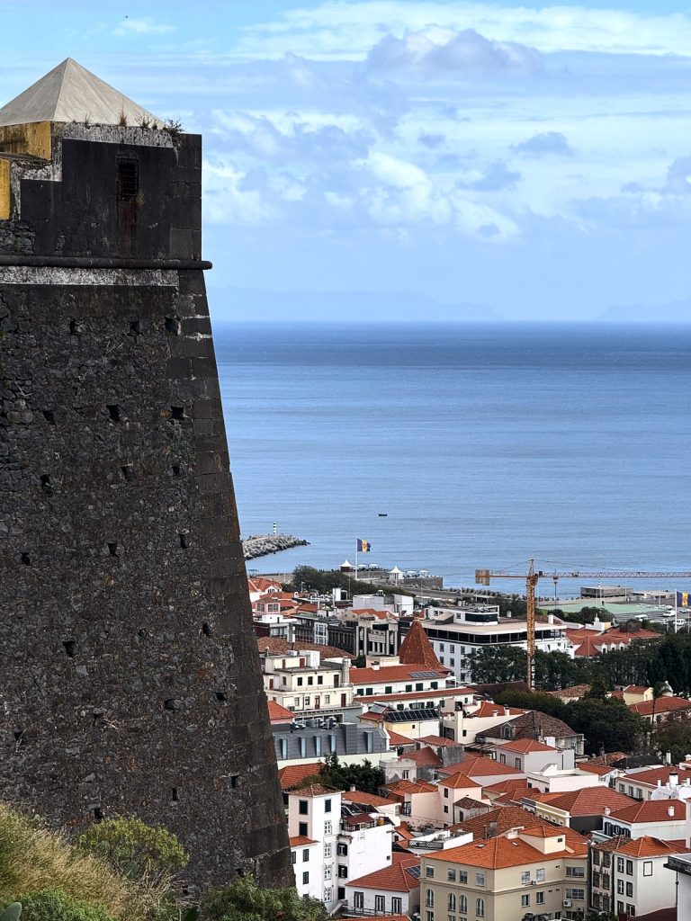 Ein dunkler steinerner Wachturm der Festung mit Pyramidendach, daneben der Blick hinunter auf Funchals rote Dächer, den Hafen und den Ozean