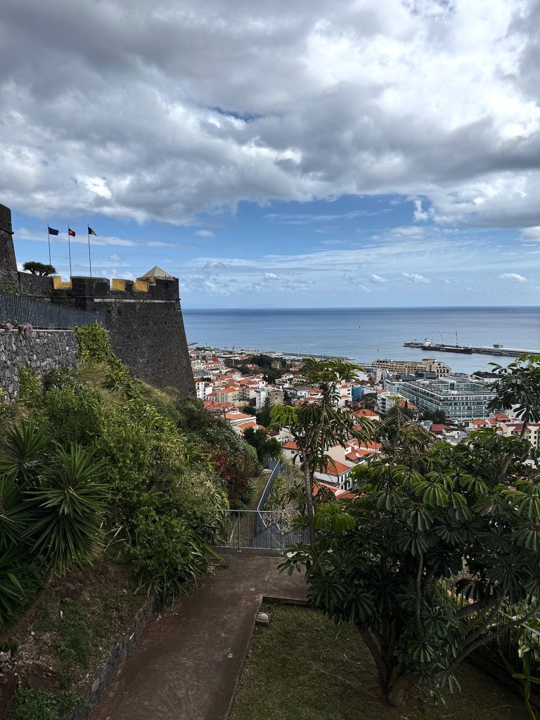 Ein Weg durchs Grün unterhalb der Mauern der Fortaleza do Pico, drei Flaggen auf den Mauern und ein Eckwachturm mit gelben Zinnen, dahinter die Dächer von Funchal, der Hafen und das Meer