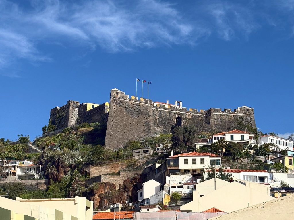 Fortaleza do Pico – die dunklen Basaltmauern der Festung mit drei Flaggen oben auf dem Hügel, darunter Häuser mit roten Dächern, darüber blauer Himmel mit Schleierwolken