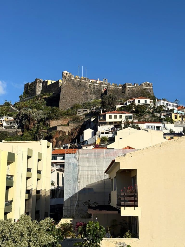 Die Steinmauern der Fortaleza do Pico mit Fahnen auf dem Hügel, darunter weiße Häuser und helle Wohnblocks im Zentrum von Funchal