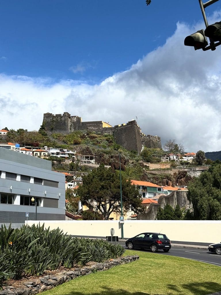 Die Mauern der Fortaleza do Pico auf einem grünen Hügel unter einer riesigen weißen Wolke, im Vordergrund ein Rasen mit Agaven, eine Straße mit Autos und ein graues Gebäude