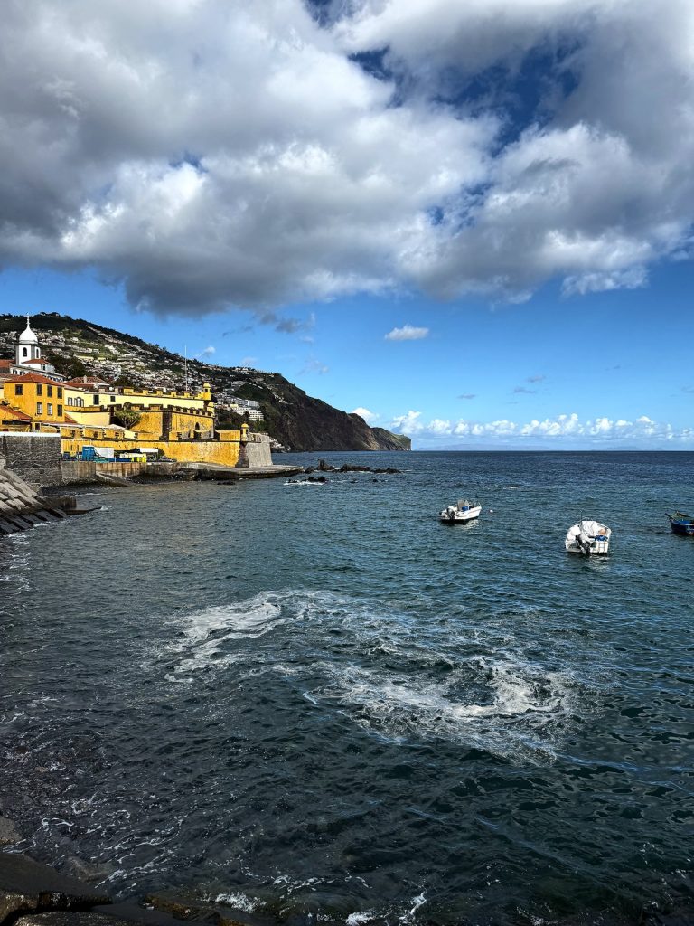 Eine Bucht mit kleinen Booten, in der Ferne die gelbe Festung São Tiago, ein weißer Kirchturm und der Häuserhang im Osten von Funchal