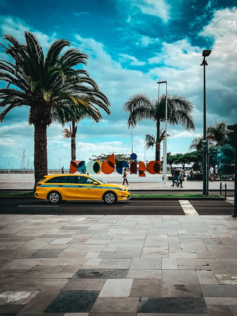 Gelbes Taxi auf der Avenida do Mar in Funchal, dahinter Palmen und der bunte Madeira-Schriftzug