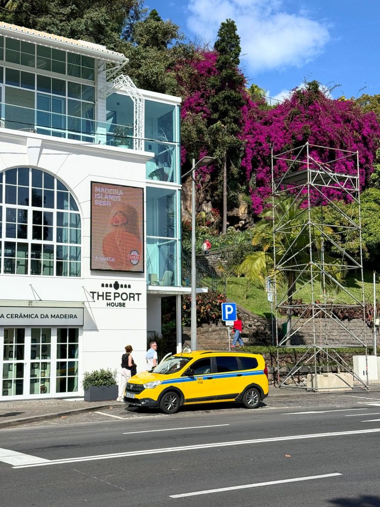 Gelbes Taxi vor einem weißen Gebäude mit großen Fenstern, daneben eine blühende lila Bougainvillea