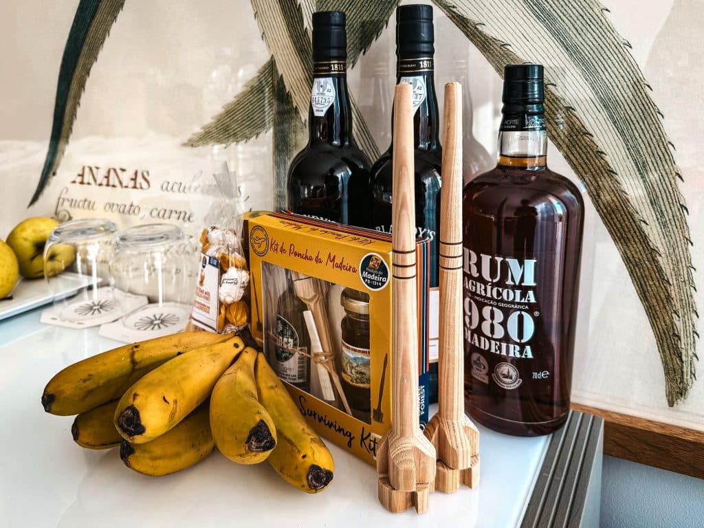 Flaschen mit Madeirawein und Rum, Bananen, Süßigkeiten und hölzerne Poncha-Quirle auf einem Tisch im Hotelzimmer