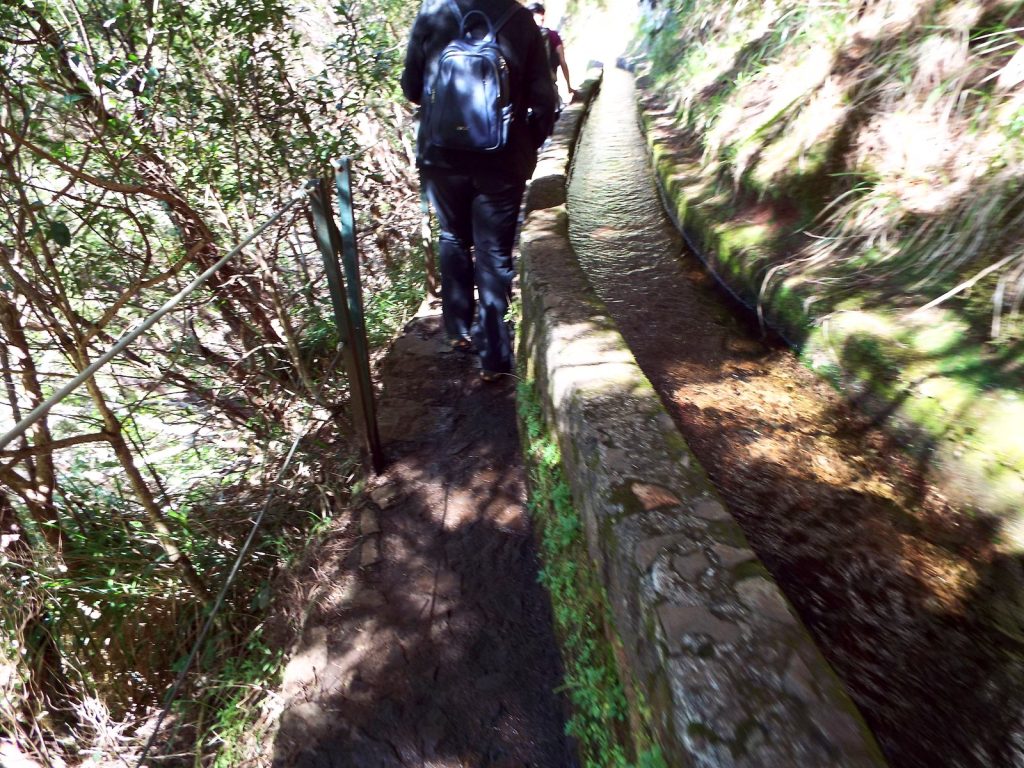 Ein Wanderer mit Rucksack, von hinten gesehen, geht auf einem schmalen Pfad neben einem Levada-Kanal mit fließendem Wasser, daneben ein Metallgeländer