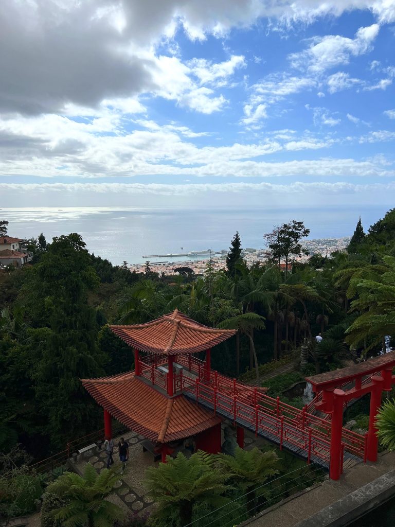 Eine rote Pagode und eine Brücke im Orientalischen Garten des Monte Palace, in der Ferne Funchal und der Ozean unter Wolken