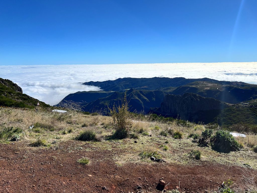Ein Wolkenmeer unterhalb der Bergkämme beim Pico do Arieiro, im Vordergrund rote Erde und trockene Sträucher