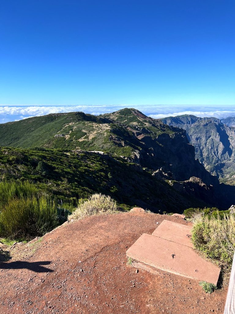 Ein roter Weg und Betonplatten an einem Aussichtspunkt, dahinter grüne Bergkämme und ein Wolkenmeer