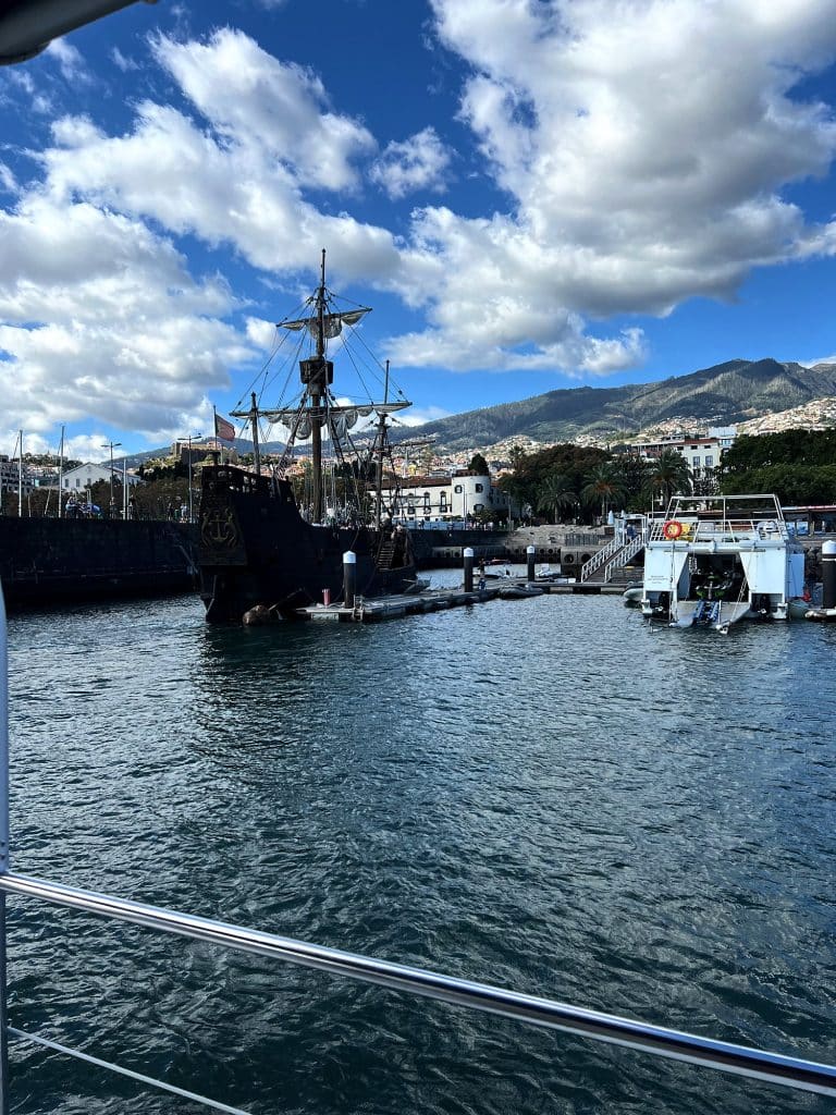 Der hölzerne Nachbau der Santa Maria am Kai im Hafen von Funchal, dahinter die Hänge der Stadt und Wolken am blauen Himmel