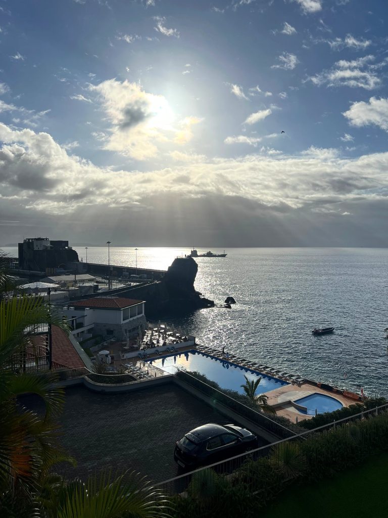 Die Sonne bricht über dem Ozean durch die Wolken, darunter Hotelpools, eine Mole und ein Felsen im Meer im Westen von Funchal