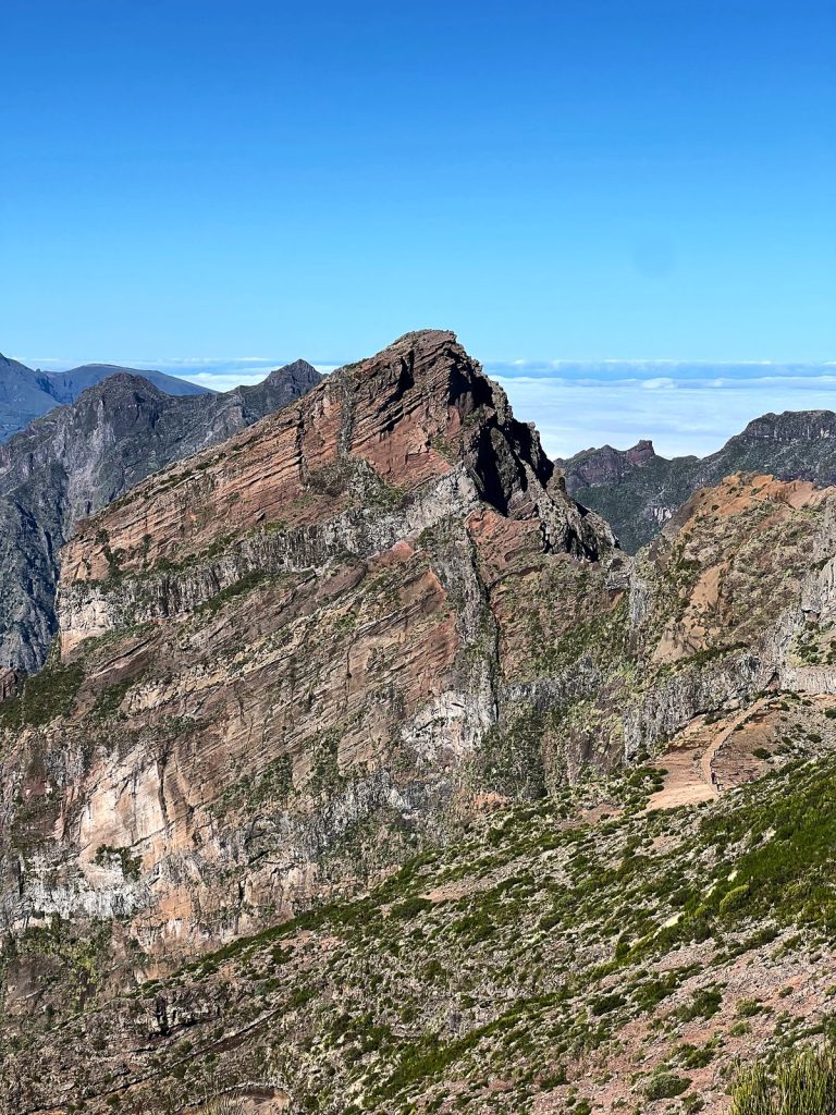 Ein rotbrauner Felsgipfel mit Schichten aus Vulkangestein, dahinter weitere Kämme und ein Wolkenmeer