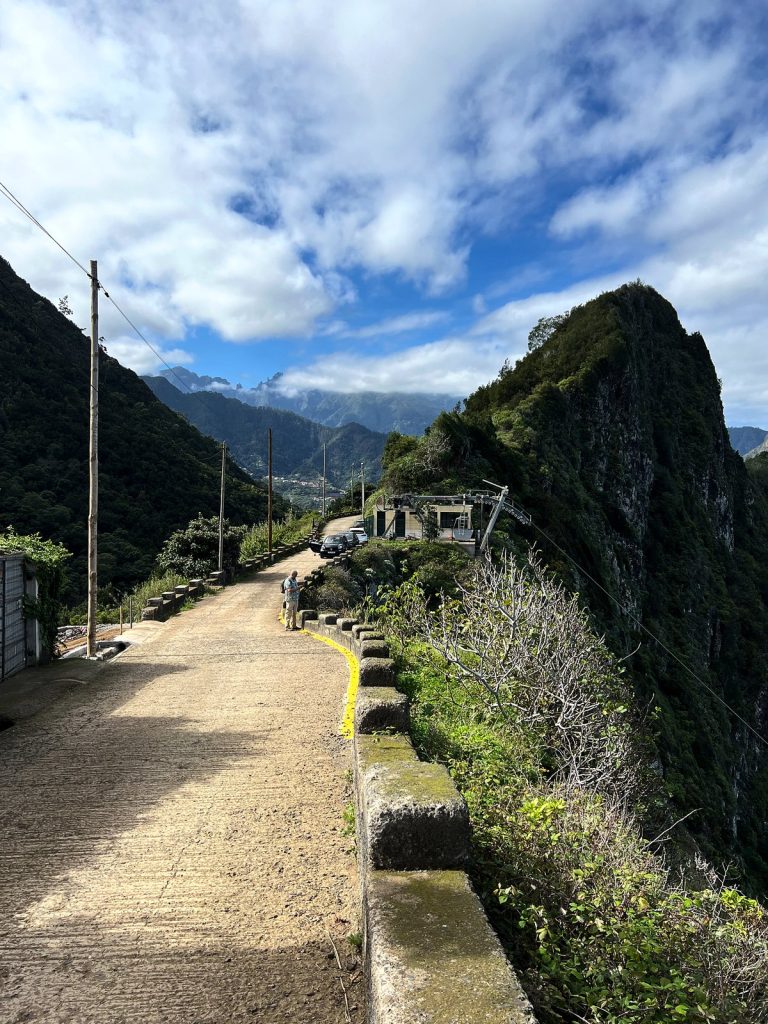 Ein schmaler Pfad entlang einer Steinmauer über einer steilen grünen Klippe, in der Ferne Berge und Wolken
