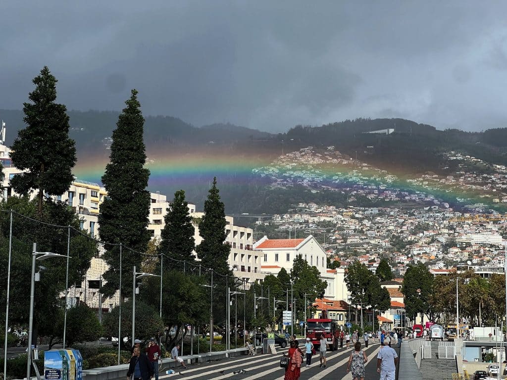 Ein Regenbogen über den Hängen von Funchal unter dunklen Wolken, darunter die baumbestandene Promenade, Gebäude und kleine Gestalten von Passanten