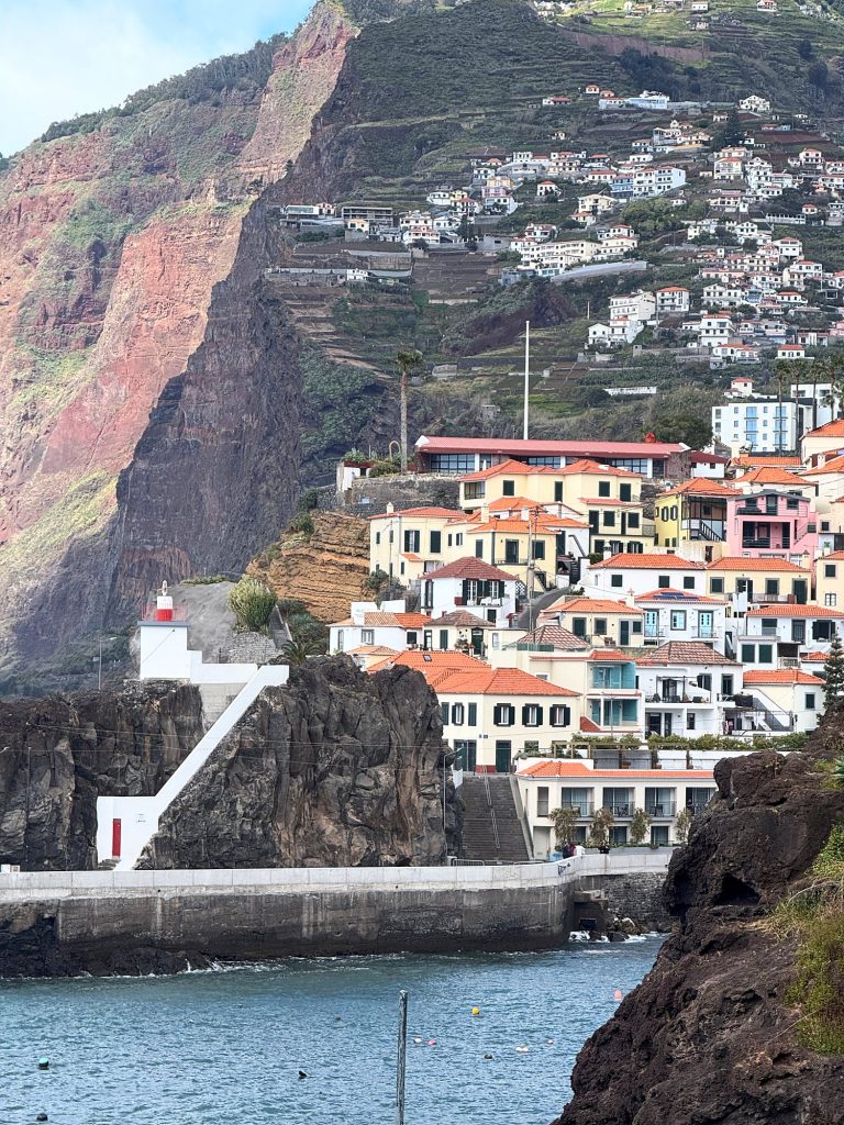 Bunte Häuser von Câmara de Lobos auf einem Felsen über dem Wasser, dahinter die rote Felswand und Terrassen am Hang