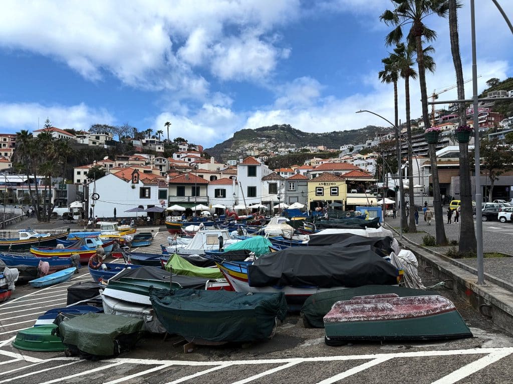 Fischerboote unter Planen auf dem Platz am Hafen von Câmara de Lobos, dahinter weiße Häuser, Palmen und ein grüner Hang