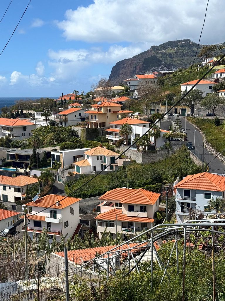 Weiße Häuser mit orangefarbenen Dächern an einem steilen Hang, dahinter die Steilküste Cabo Girão unter Wolken
