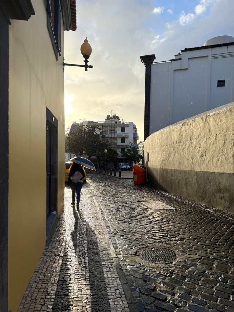 Eine schmale, nach Regen nasse Pflastergasse in Funchal, ein Passant mit Schirm geht ins Licht, rechts eine weiße Mauer