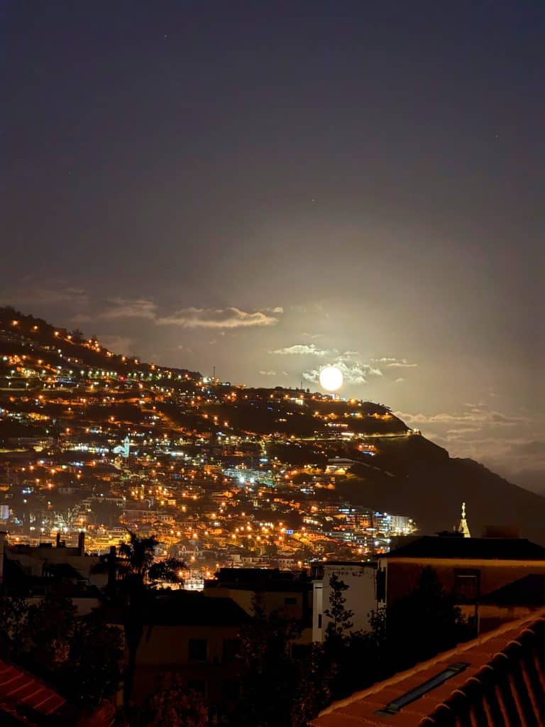 Ein Vollmond tief über dem beleuchteten Hang von Funchal bei Nacht, vorne Dächer
