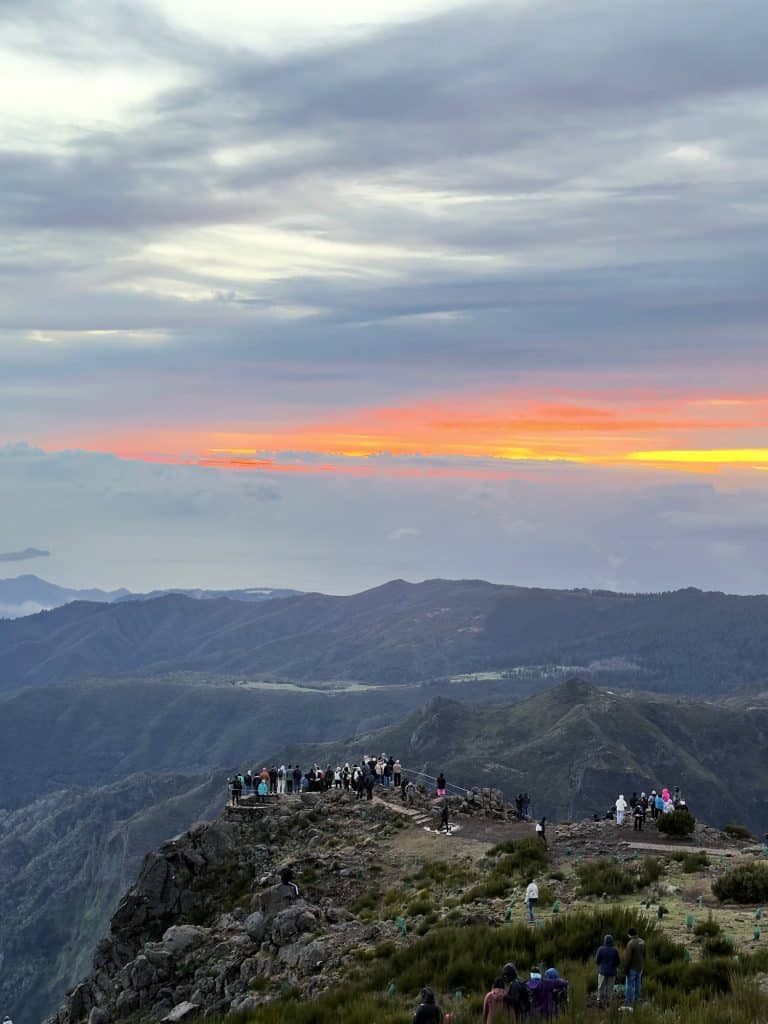 Menschen auf einem felsigen Aussichtspunkt am Pico do Arieiro in der Morgendämmerung, darüber ein orangefarbener Himmelsstreifen über den Bergkämmen
