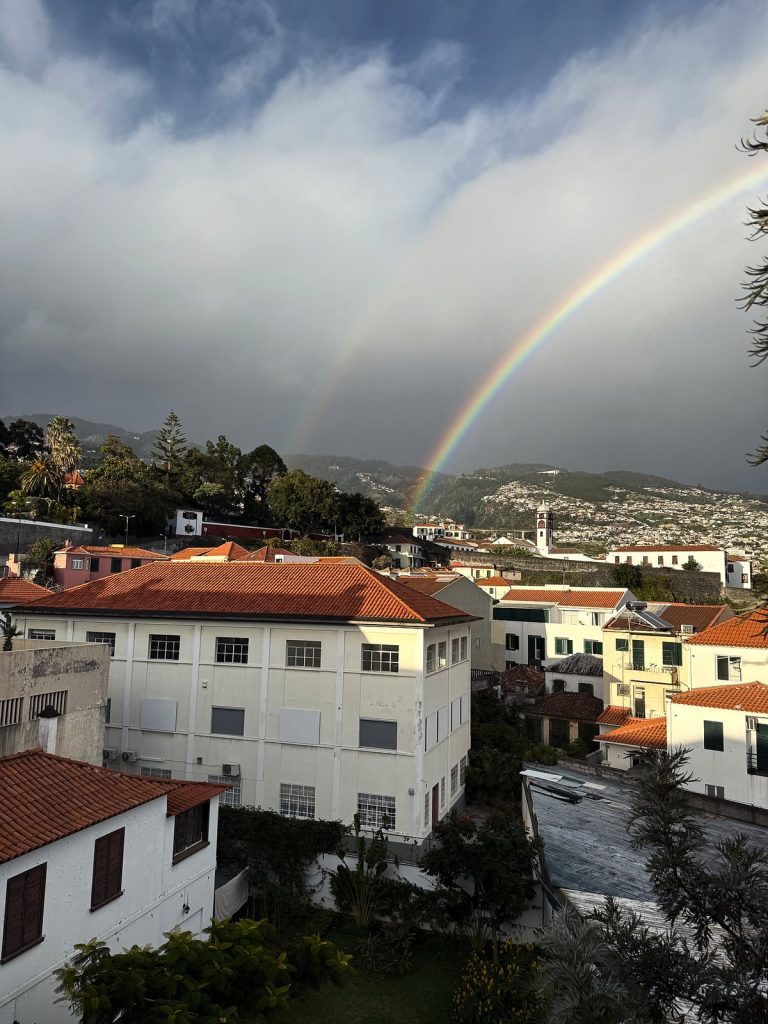 Ein doppelter Regenbogen über den Dächern von Funchal unter dunklen Regenwolken, vorne ein weißes Gebäude mit orangefarbenem Dach, dahinter Häuser an den Hängen