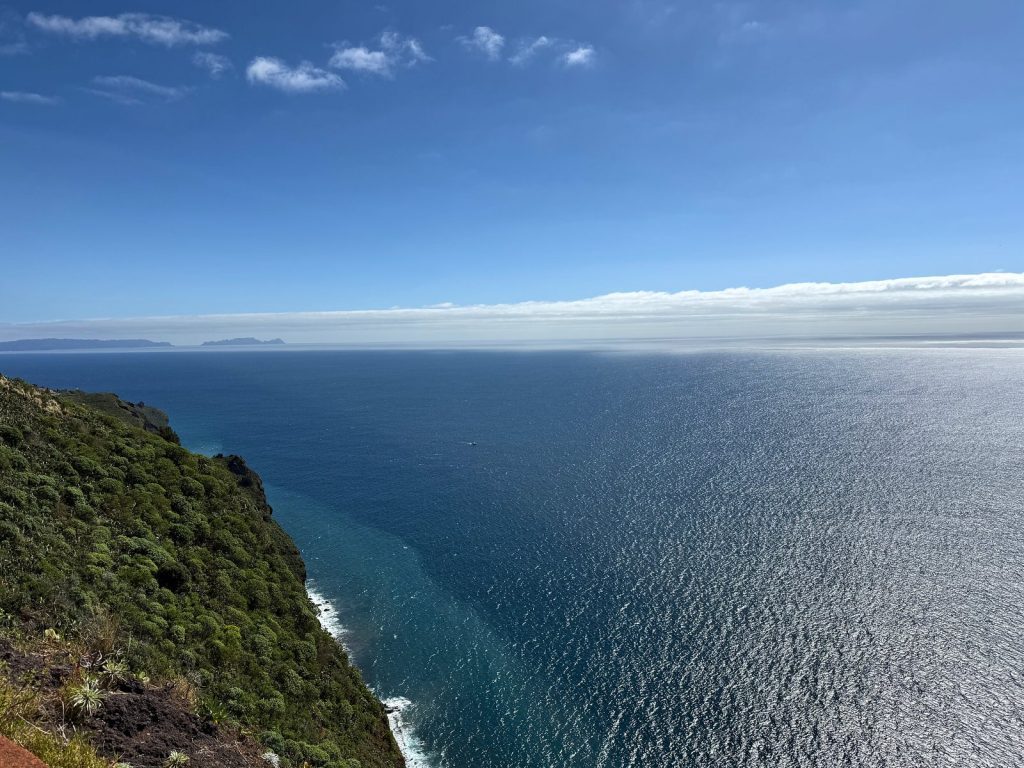 Ein grüner Klippenhang fällt in den Ozean ab, am Horizont ein Wolkenband