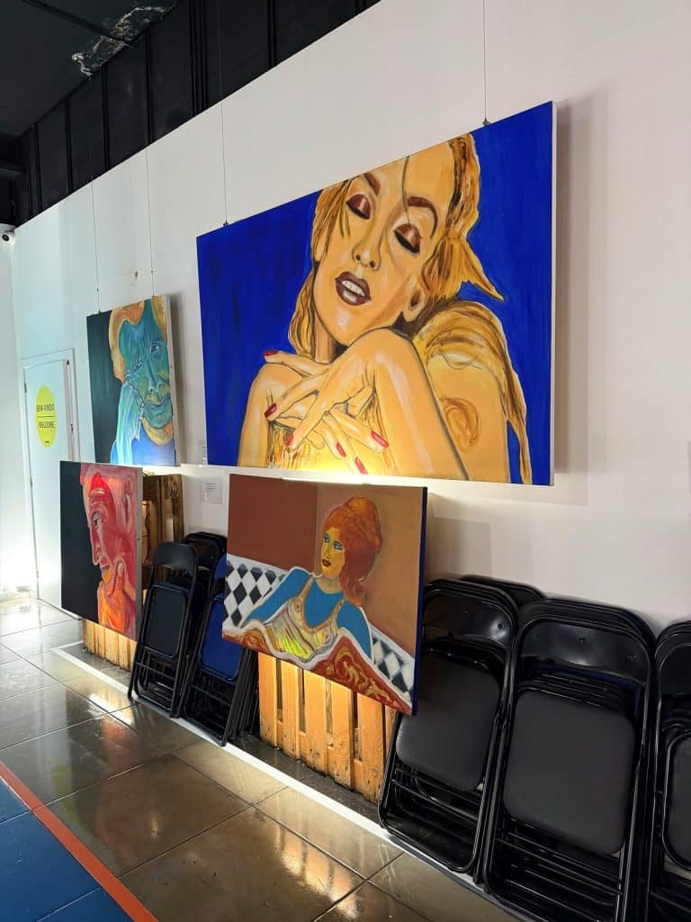 Große bunte Pop-Art-Gemälde an der weißen Wand eines Flurs, darunter angelehnte Leinwände und zusammengeklappte Stühle