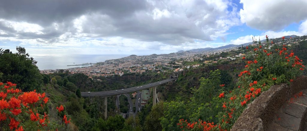 Ein weites Panorama über Funchal und den Ozean von einer Gartenterrasse, unten ein Tal mit Viadukt, im Vordergrund Sträucher mit roten Blüten