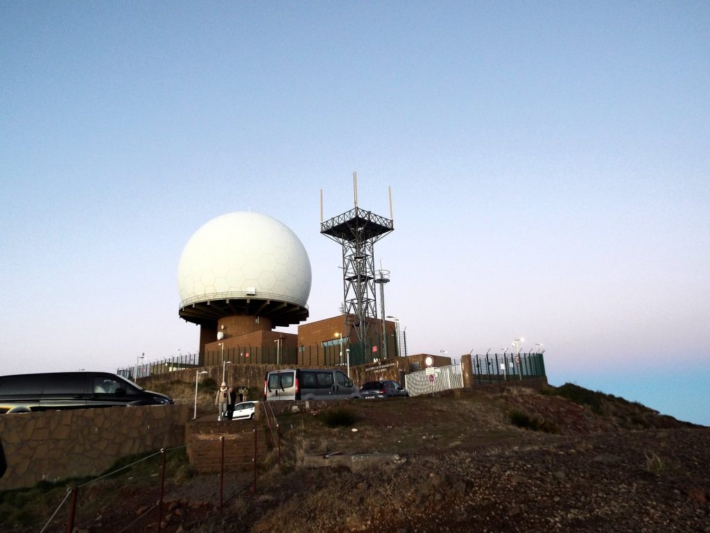 Die Radarkuppel und ein Stahlmast mit Antennen auf dem Gipfel des Pico do Arieiro, geparkte Autos an den Gebäuden, rosa Himmel in der Morgendämmerung