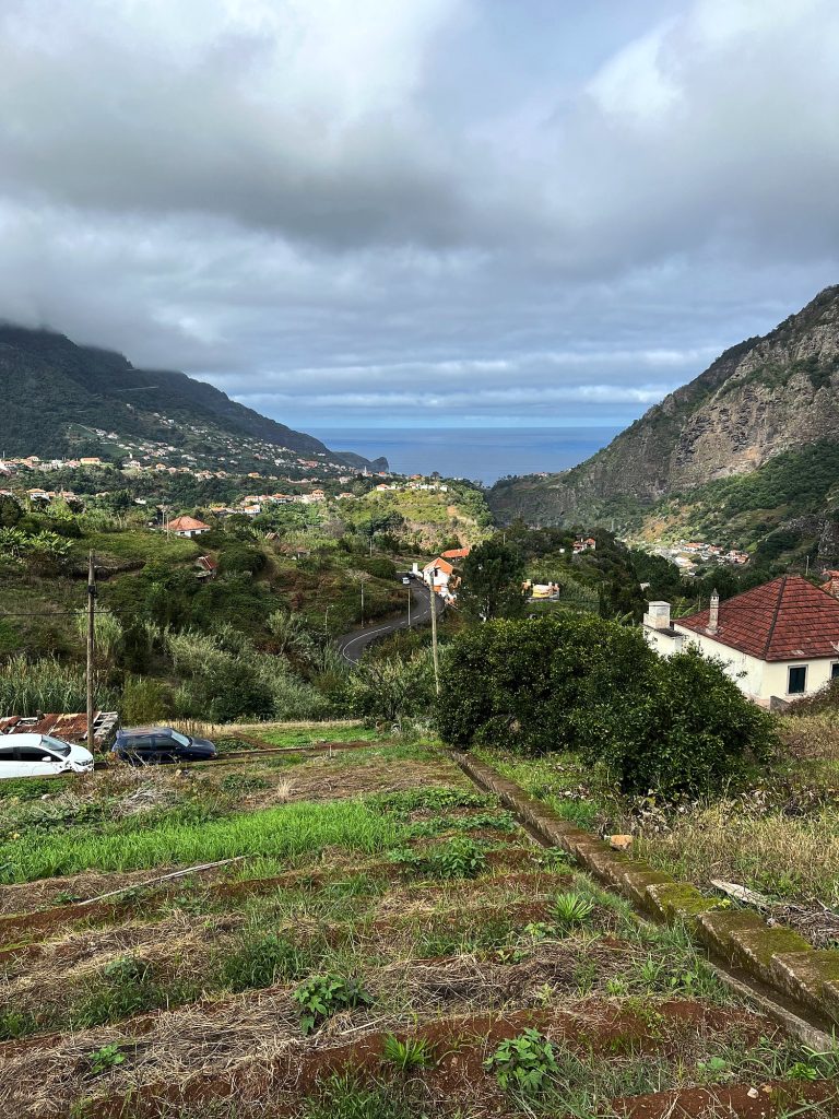 Weites grünes Tal mit Feldern und Häusern, das zwischen wolkenverhangenen Bergen zum Meer abfällt