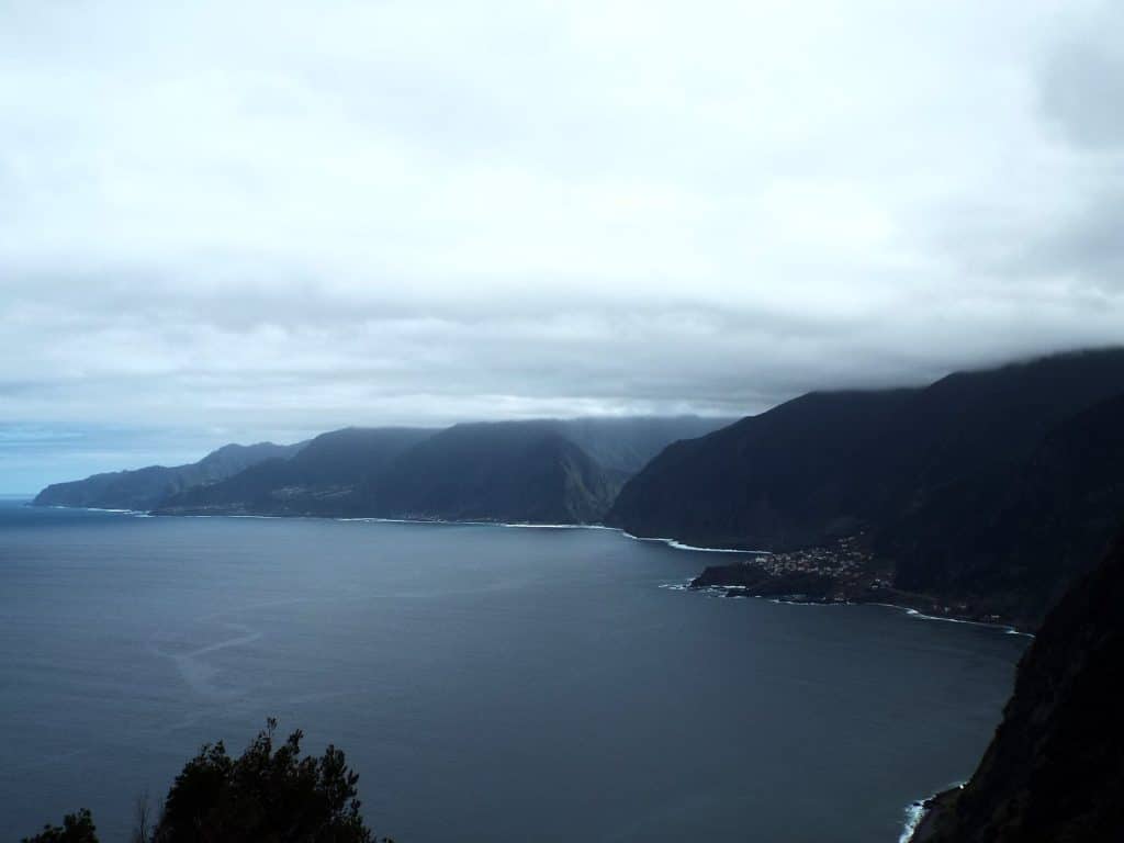 Bedeckter Himmel über der Nordküste Madeiras, dunkle Klippen fallen in einen grauen Ozean ab, Wolken verdecken die Gipfel, am Ufer ein kleines Dorf