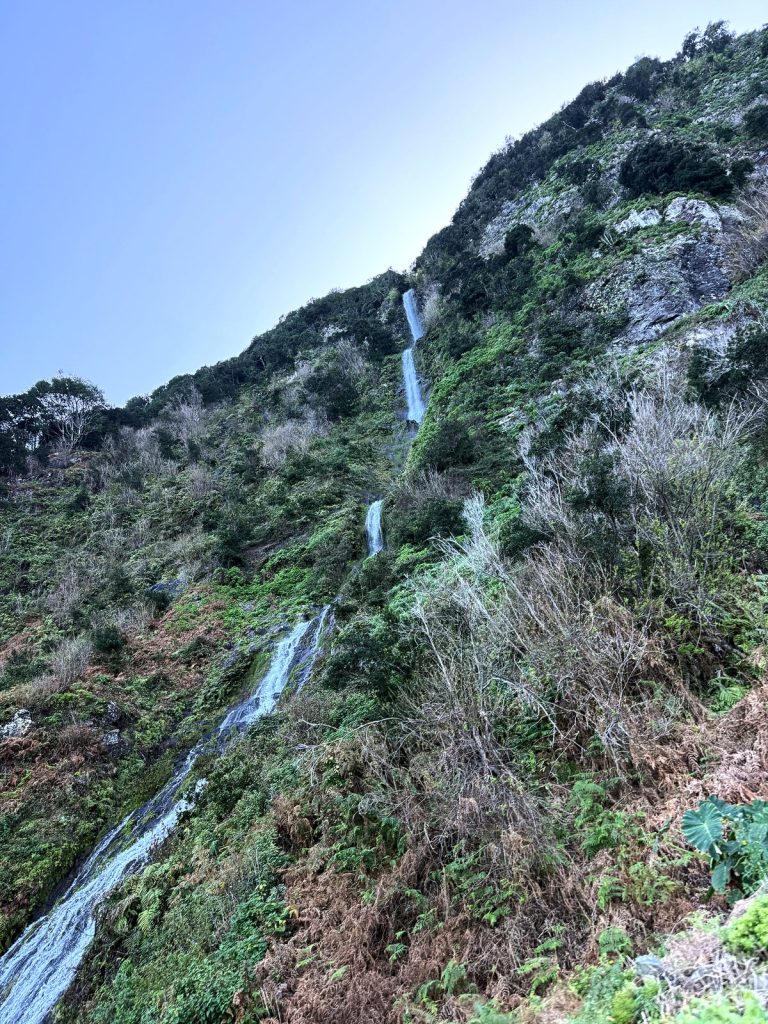 Ein schmaler Wasserfall stürzt in Kaskaden einen steilen, mit Buschwerk bewachsenen Hang hinunter, darüber blauer Himmel